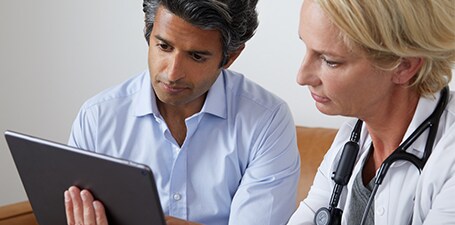 Arzt, der auskultiert und dem Mann die App auf dem Tablet zeigt
